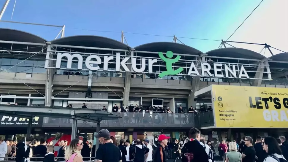 Foto in Beitrag von 5min.at: Zu sehen ist die Merkur Arena bei schönem Wetter und blauem Himmel. Viele Menschen warten davor auf den Einlass.