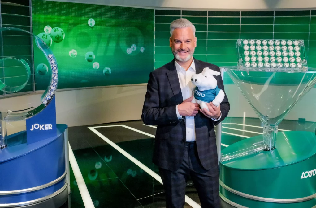 Foto in Beitrag von 5min.at: Zu sehen ist einer der drei Lottomoderatoren im Studio. Er hat ein Pl&uuml;schtier in den Armen.