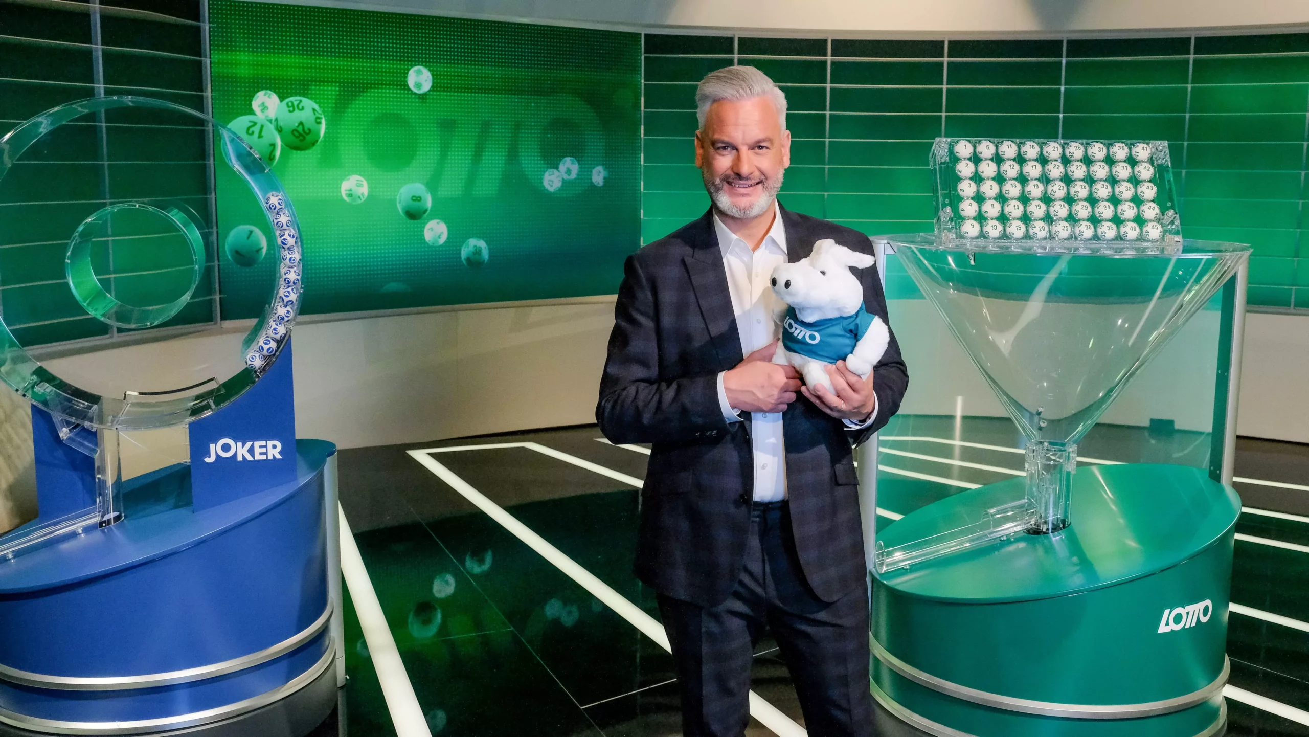 Foto in Beitrag von 5min.at: Zu sehen ist einer der drei Lottomoderatoren im Studio. Er hat ein Plüschtier in den Armen.