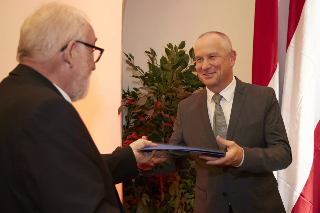 Zu sehen sind zwei Männer. Der eine übergibt dem anderen ein Formular