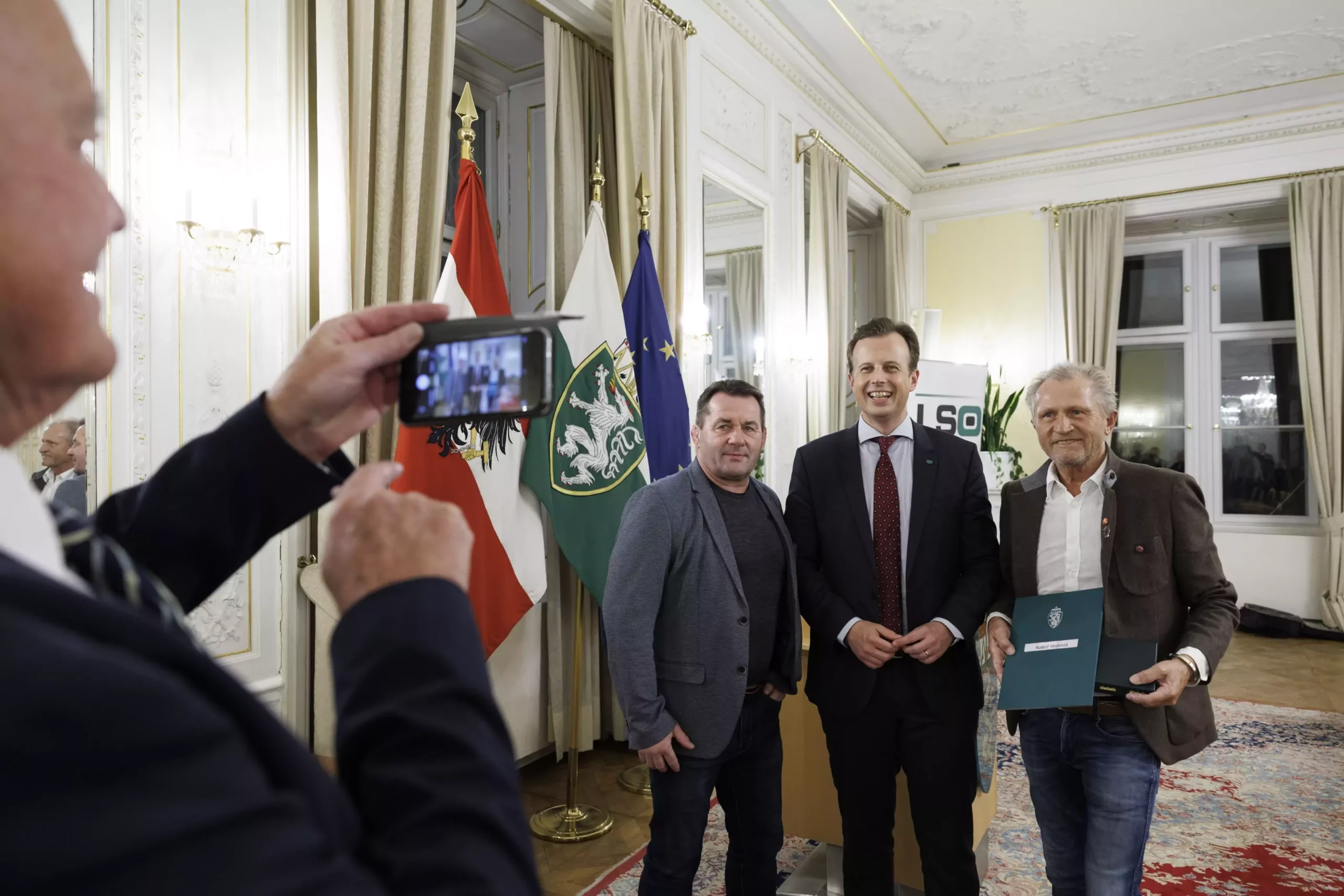 Das Bild auf 5min.at zeigt: Mehrere Personen stehen im Weißen Saal der Grazer Burg nebeneinander und werden durch ein Smartphone fotografiert.