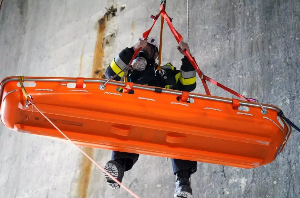 Ein Bild auf 5min.at zeigt einen Feuerwehrmann, der gerade eine orange Trage abseilt. Beide hängen in der Luft, dahinter ist eine graue Betonwand.