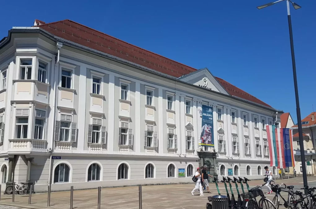 Ein Bild auf 5min.at zeigt die Außenfassade des Klagenfurter Rathauses, davor schlendern Spaziergänger durch die Stadt. Der Himmel ist blau.