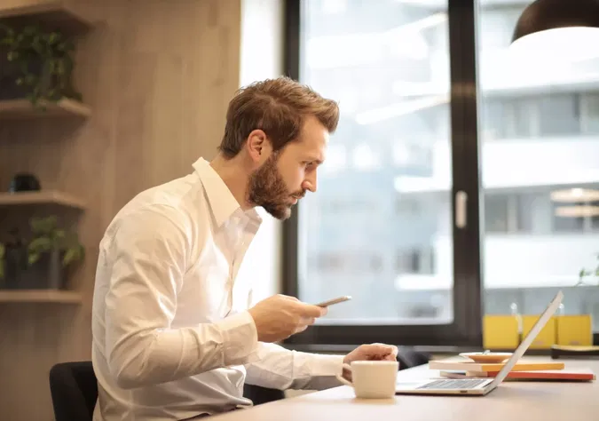 Ein Bild auf 5min.at zeigt einen formal gekleideten Mann, der an einem Laptop sitzt. Neben ihm steht eine Tasse Kaffee. In seiner Hand hält er ein Smartphone.