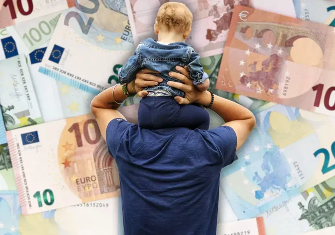 Foto in Beitrag von 5min.at: Zu sehen ist ein Vater mit einem Kind, im Hintergrund leicht verschwommen Geld.