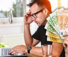 Foto in Beitrag von 5min.at zeigt im Vordergrund eine Hand mit mehreren Euroscheinen, im Hintergrund eine Person vor einem Laptop, der angestrengt auf den Bildschirm schaut.