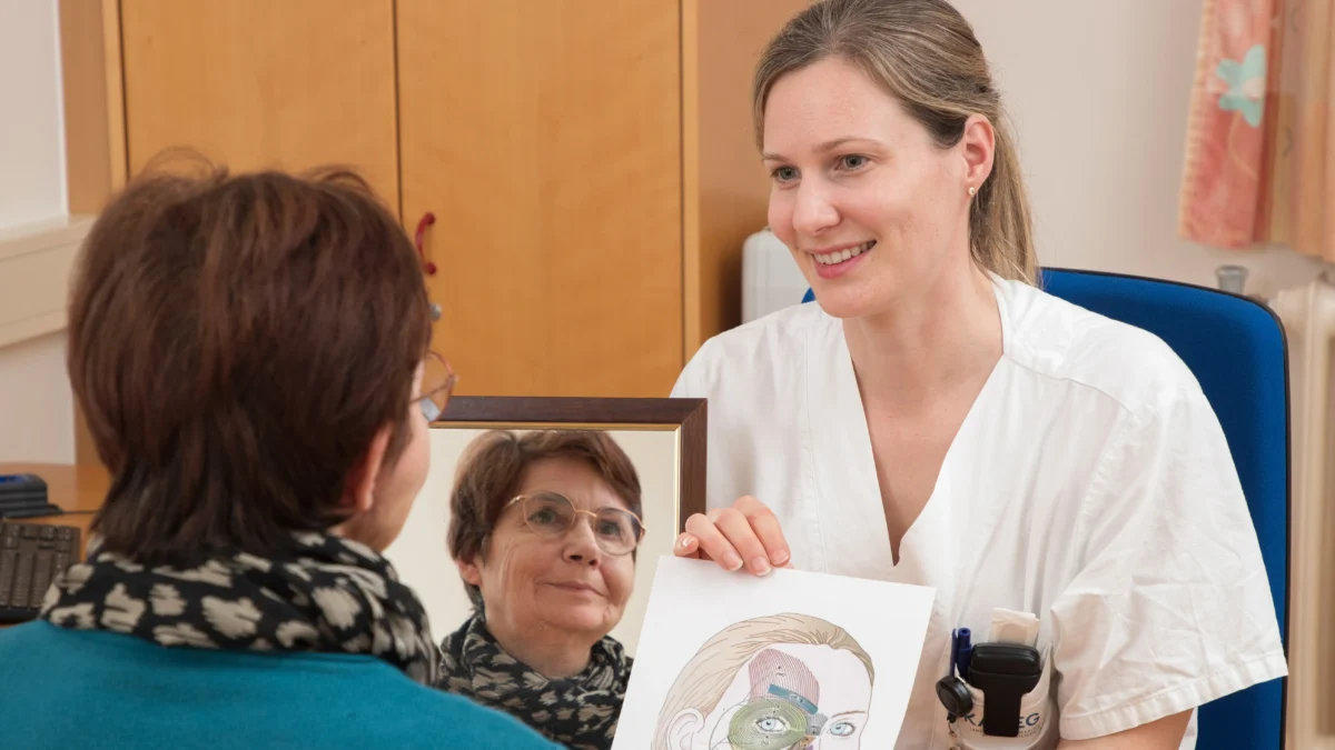 Ein Bild auf 5min.at zeigt eine Patientin, die in einen Spiegel lächelt. Neben ihr sitzt eine Ärztin.