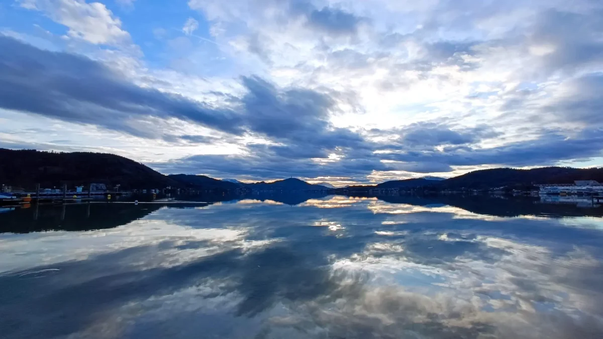 Ein Bild auf 5min.at zeigt den Wörthersee. Über den Himmel ziehen Wolken, diese spiegeln sich auch im Wasser.