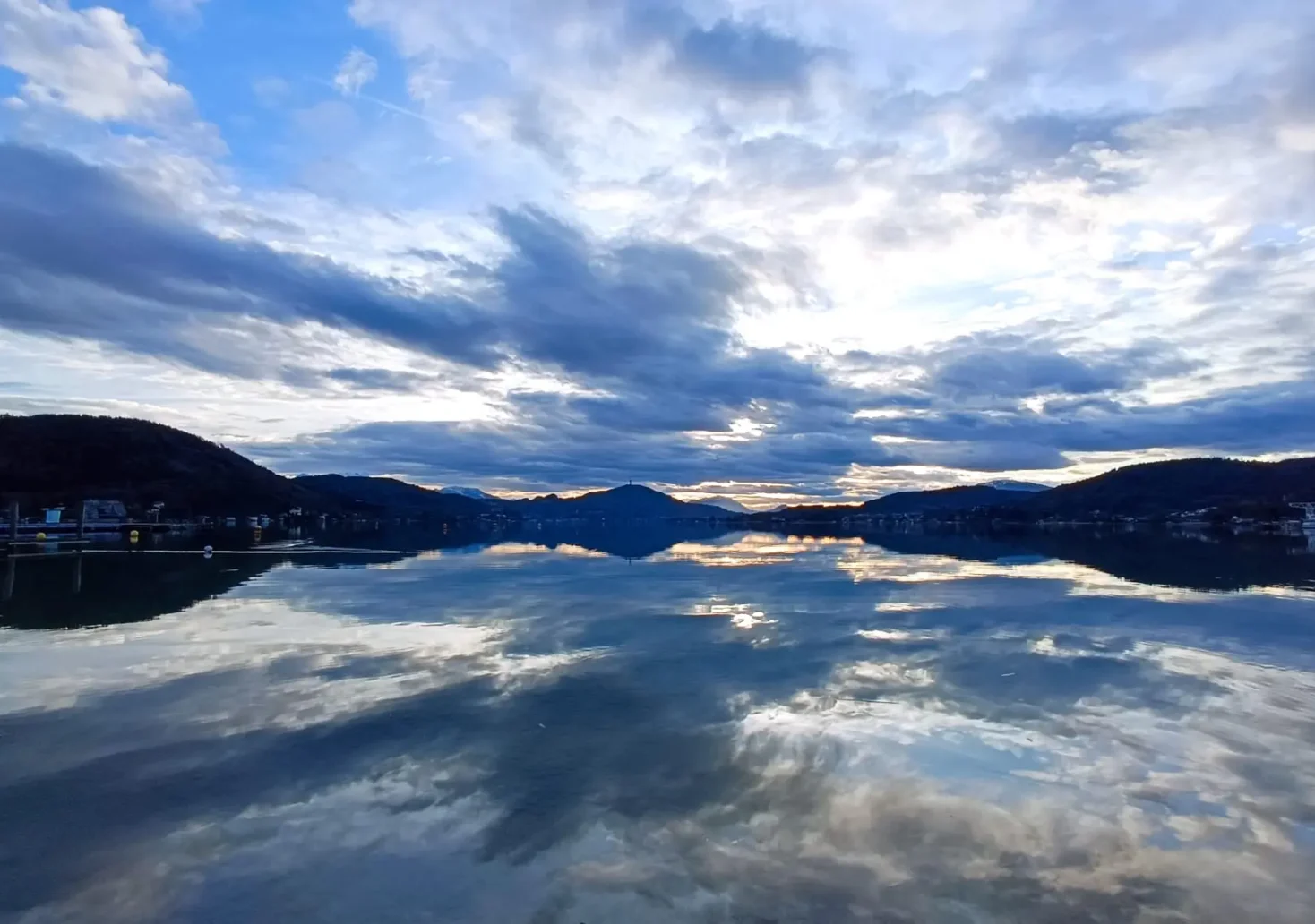 Ein Bild auf 5min.at zeigt den Wörthersee. Über den Himmel ziehen Wolken, diese spiegeln sich auch im Wasser.