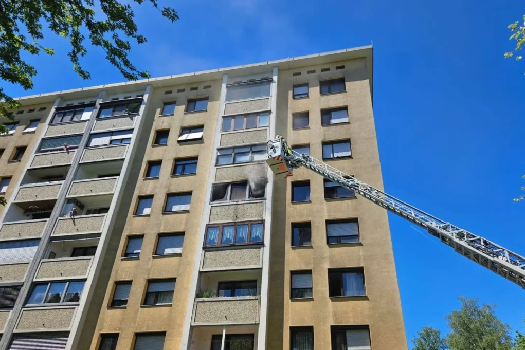 Ein Bild auf 5min.at zeigt, wie Rauch aus der Wohnung eines Mehrparteienhauses aufsteigt. Die Drehleiter ist davor positioniert.